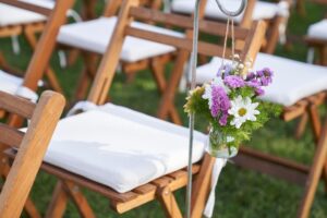 drewniane krzesła z bialymi poduszkami na wesele w ogrodzie z kwiatkami na poręczach(1)